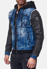 Veste en denim et cuir avec un corps en denim bleu usé, des manches en cuir noir, des poches avant et un patch logo sur l'épaule.