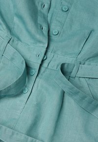 Una prenda abotonada de color azul verdoso hecha de tela texturizada, con botones, una cintura con cinturón y un cuello estructurado.