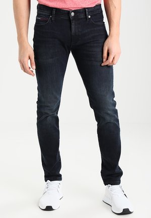 Man draagt slanke donkerblauwe jeans, witte sneakers en een rood shirt, staand tegen een effen lichte achtergrond.