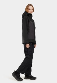 Black waterproof jacket with a grey panel, hood, and zippered pockets paired with cargo pants and black sneakers. Smooth texture, relaxed fit.