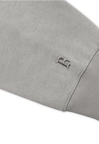 Primo piano di una manica in tessuto grigio chiaro con polsino a coste e piccolo logo ricamato "RG" vicino alla cucitura.