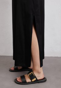 Robe en satin noir avec une fente latérale, associée à des sandales plates noires ornées d'une boucle dorée et de coutures blanches sur la semelle.