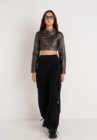 Μεταλλική γκρι cropped δερμάτινη μπουφάν με ψηλό γιακά, λεπτομέρειες με κουμπιά, συνδυασμένη με μαύρο παντελόνι cargo και μαύρα δερμάτινα παπούτσια με κορδόνια.