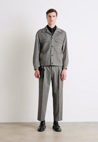 Grauer, gewebter Anzug mit einer Knopfjacke, zwei Fronttaschen und geraden Hosenbeinen. Das Model hält eine schwarze Clutch und trägt schwarze Schuhe.