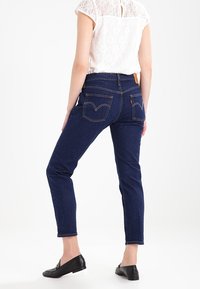 Jeans in denim blu scuro con una vestibilità slim, cinque tasche e dettagli di cucitura sottili. Indossati con un top in pizzo chiaro e scarpe nere.