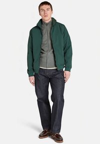Chaqueta bomber verde sobre una sudadera con capucha gris con cremallera, combinada con jeans oscuros de mezclilla y zapatos marrones. El modelo está de pie, con las manos relajadas.