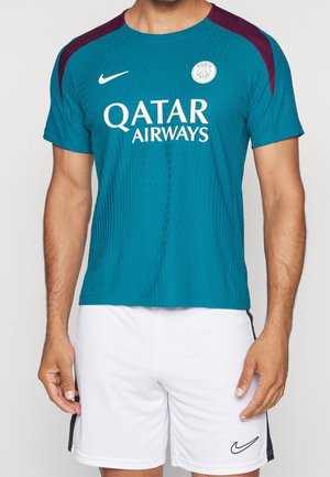 Kolorowa na niebiesko-zielono koszulka piłkarska Paris Saint-Germain z logo Qatar Airways, noszona z białymi szortami Nike, na stojącej osobie pokazanej od szyi do połowy uda.