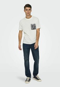 T-shirt bianca in cotone con tasca sul petto, decorata con un motivo a foglie in bianco e nero, abbinata a jeans blu scuro e sneakers blu navy.