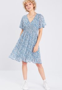 Robe florale bleue avec des manches courtes en volants, un corsage smocké et une jupe fluide. Présente un décolleté en V et des motifs floraux blancs répartis sur l'ensemble.