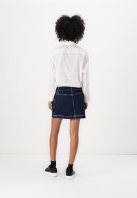 Hvit skjorte med diskret logomønster, kombinert med en mørk denimskjørt. Outfiten fullføres med svarte sneakers og ankelstrømper.