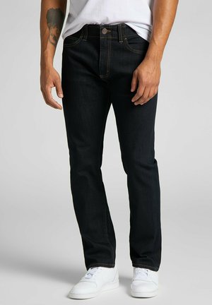 Dark blue denim jeans with a slim fit, visible stitching in contrasting gold, and five-pocket design. Worn with a white T-shirt and sneakers.