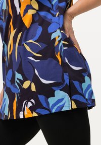 Bluse mit Blumenmuster, die lebendige blaue, orange und gelbe Blätter auf einem dunklen Hintergrund zeigt. Verfügt über seitliche Schlitze am Saum für mehr Bewegungsfreiheit.