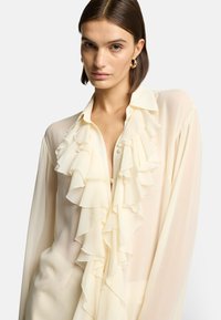 Blusa crema con dettagli a volant nella parte anteriore, tessuto morbido e trasparente, colletto con bottoni e maniche lunghe, presentata su uno sfondo neutro.
