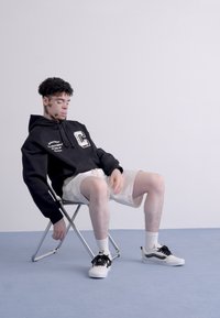 Felpa nera con scritte bianche, pantaloni corti chiari, seduto su una sedia pieghevole, indossando sneakers nere e bianche su un pavimento blu.