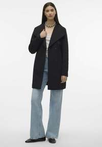 Svart dubbelknäppt rock med bred krage, i kombination med ljusblå utsvängda jeans och svarta skor. Har en mjuk tygtextur.