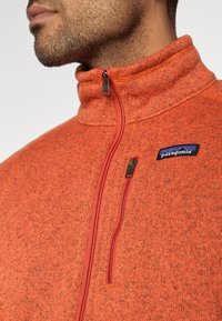 Πλειοφόρεμα από πορτοκαλί φλις με φερμουάρ μέχρι τη μέση, υφασμένο με υφή, κόκκινο φερμο υάρ και μπλε πατσηόν με το λογότυπο της Patagonia στο στήθος. Κοντινή όψη.