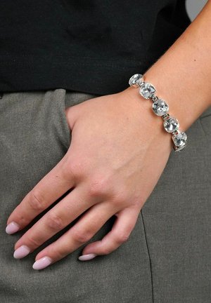 Silverarmband med flera klara ädelstenar i en länkad kedjedesign, bärs på en hand som vilar mot en grå tygbakgrund.