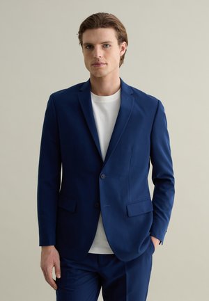 Chaqueta de traje azul marino con dos botones, solapas de muesca y bolsillos laterales. Lleva puesta una camiseta blanca de cuello redondo. Textura suave, corte entallado.