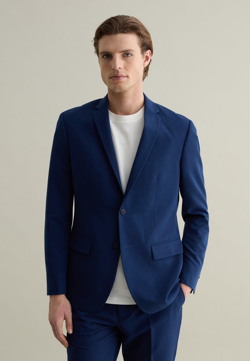 Giacca da abito blu navale con due bottoni, revers a punta e tasche laterali. Indossata su una t-shirt bianca a girocollo. Texture liscia, vestibilità su misura.