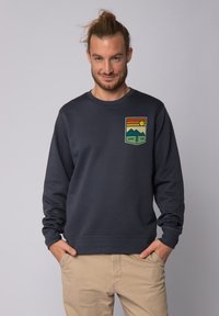 Marineblauwe sweat met geribbelde manchetten en tailleband, met een kleurrijke grafische patch van bergen en de zon met de tekst "GOOD VIBE."