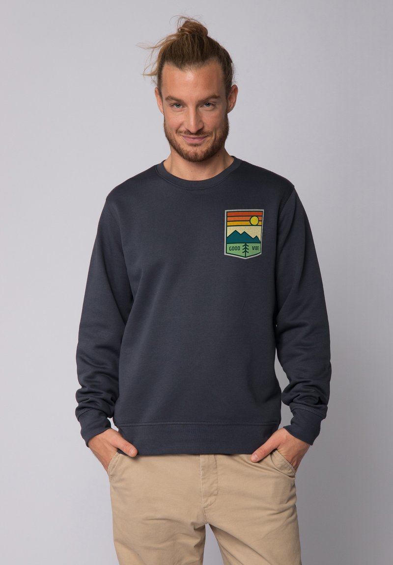 Marineblauwe sweat met geribbelde manchetten en tailleband, met een kleurrijke grafische patch van bergen en de zon met de tekst "GOOD VIBE."