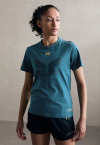 Türkis puuvillane T-särk musta silmadega mustri ja kuldse logoga rinnal. Lühikesed varrukad ja klassikaline lõige. Kombineeritud mustade lühikesed püksidega.