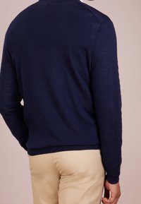 Navy blue knitted sweater with a round neck and ribbed hems, featuring a smooth texture and set-in sleeves, paired with beige trousers.