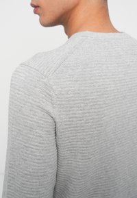 Άνδρας φορώντας ένα ανοιχτό γκρι πλεχτό πουλόβερ, απεικονίζεται από πίσω και ελαφρώς στραμμένος στο πλάι.