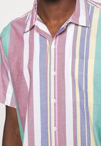 Primo piano di una persona che indossa una camicia a maniche corte con bottoni, a righe verticali in rosa, bianco, blu, giallo e verde, con taschino sul petto.