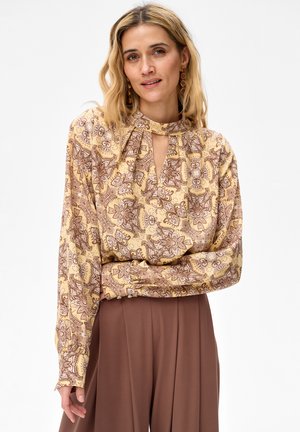 Vrouw met schouderlang blond haar, gekleed in een gele paisleyblouse met een sleutelgat aan de borst en een bruine broek met hoge taille.