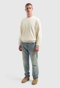 Jonge man staat in een crèmekleurige longsleeve crewneck sweatshirt, lichtblauwe distressed jeans en tan kleurige veterschoenen op een grijze achtergrond.