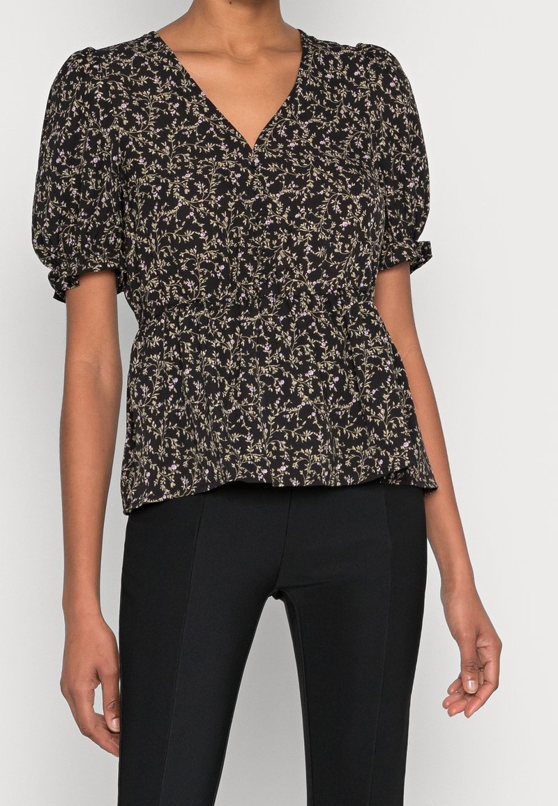 Blouse noire à motifs floraux avec un col en V, des manches bouffantes et une taille élastique. Présente de petites fleurs violettes et blanches sur un tissu doux.