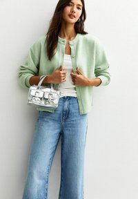 Cardigan lavorato a maglia verde chiaro con collo rotondo e chiusura con bottoni, abbinato a jeans azzurri a vita alta e una mini borsa trasparente.