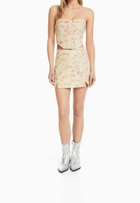 Dvoudílný outfit s květinovým vzorem, obsahující korzetový top bez ramínek v jemně žluté barvě a odpovídající mini sukni s bočními rozparky. Stříbrné kotníkové boty.