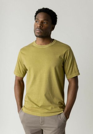 Muž s krátkymi kučeravými vlasmi, oblečený v horčicovo žltom tričku a béžových nohaviciach, stojaci s rukami v vreckách proti jednoliatej svetlej stene.