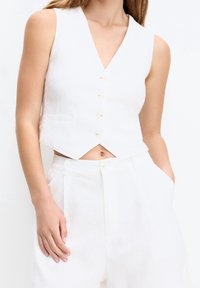 Gilet court sans manches en lin blanc avec un décolleté en V, doté de cinq boutons et de deux poches latérales. Texture lisse et design épuré.