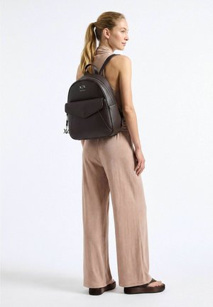 Femme avec une queue de cheval blonde portant une combinaison beige sans manches et des sandales marron, portant un sac à dos en cuir noir avec une poche avant.