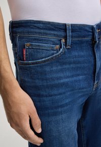 Blue denim jeans with a straight fit, featuring copper rivets, a small red tag, and a textured surface. The waistband has a slight stretch.