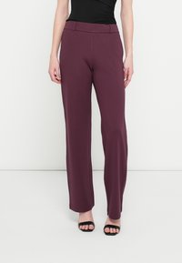 Femme portant un pantalon prune taille haute à jambe droite associé à des sandales noires à bout ouvert, debout devant un fond blanc uni.
