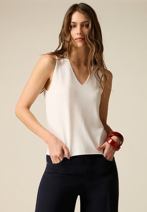 Femme aux cheveux longs portant un haut blanc sans manches à col en V, un pantalon foncé, et de larges bracelets rouges au poignet gauche, les mains dans les poches.