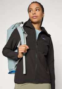 Young woman wearing a black zip-up jacket and carrying a light blue backpack, looking slightly to the side on a plain background.