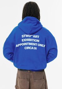 Felpa blu con testo bianco sul retro che recita "STWD™ ART EXHIBITION APPOINTMENT ONLY CIRCA 91." Design con tasca a marsupio e cappuccio.