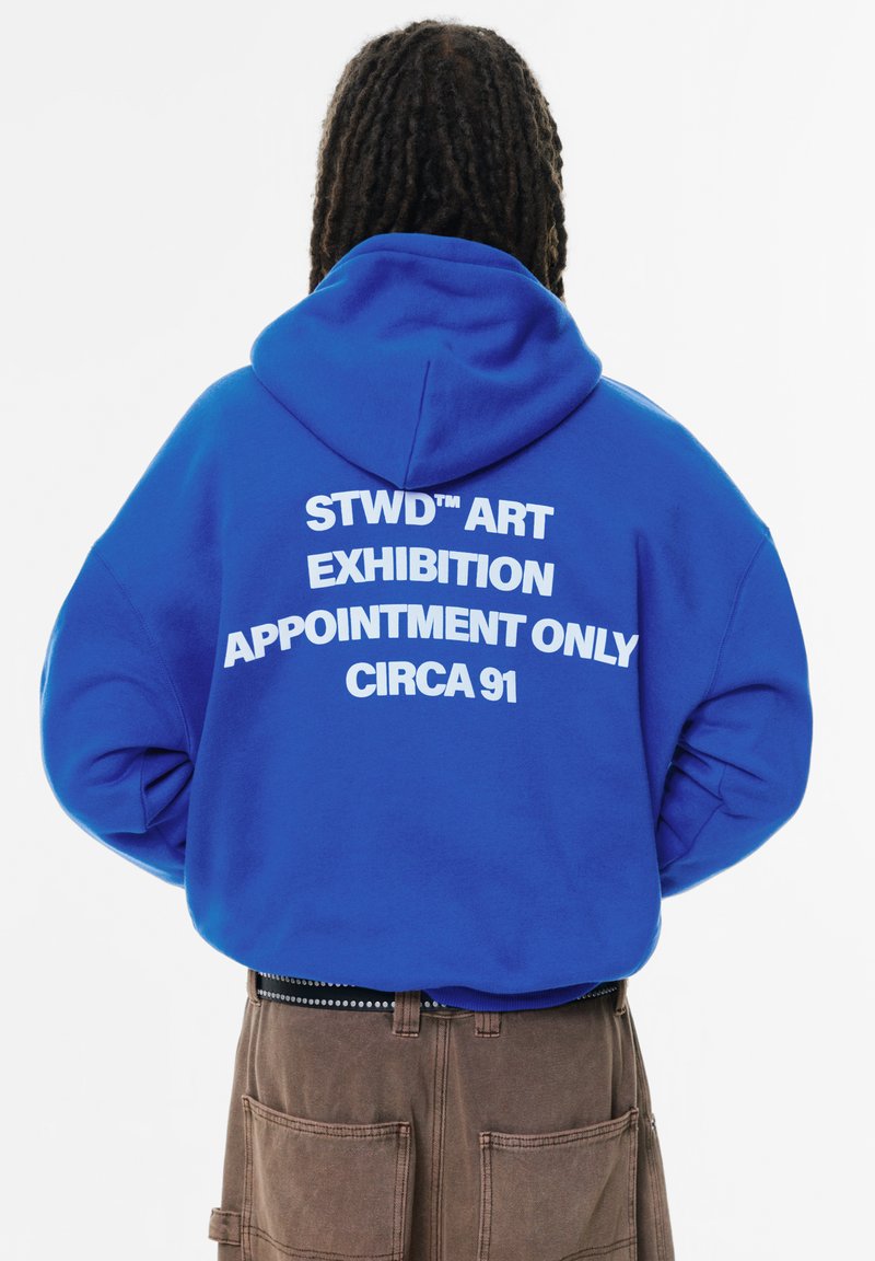 Felpa blu con testo bianco sul retro che recita "STWD™ ART EXHIBITION APPOINTMENT ONLY CIRCA 91." Design con tasca a marsupio e cappuccio.