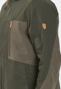 Olivgrön jacka med paneldesign som inkluderar mörkare gröna och bruna sektioner. Innehåller en dragkedjeficka och läderdetalj på axeln.