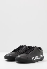 Un paio di sneakers nere basse con lacci neri e la parola "FLAWLESS" in bianco sui lati esterni su uno sfondo bianco.