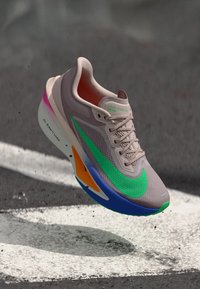 Leichtgewichtiger Laufschuh mit einem beigen Mesh-Obermaterial, grünen Akzenten, einem rosa Farbverlauf an der Ferse und einer blau-orangefarbenen Sohle.