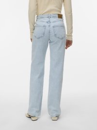 Henkilö, jolla on vaaleansiniset suorat farkut ja kermaisenvärinen pitkähihaisen yläosa, seisoo selin kameraan valkoista taustaa vasten.