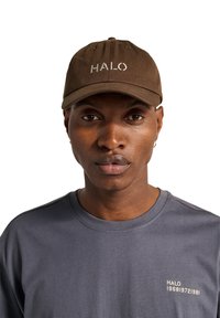 Brown cotton cap with "HALO" embroidered in white. Paired with a gray t-shirt featuring a small logo in gold near the hem.