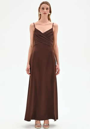 Femme portant une robe longue marron foncé sans manches avec de fines bretelles et un corsage drapé, debout devant un fond blanc uni.