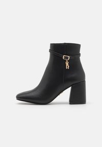 Bottine noire en simili cuir lisse, présentant un bout pointu, un talon bloc et une sangle décorative dorée avec un charm.